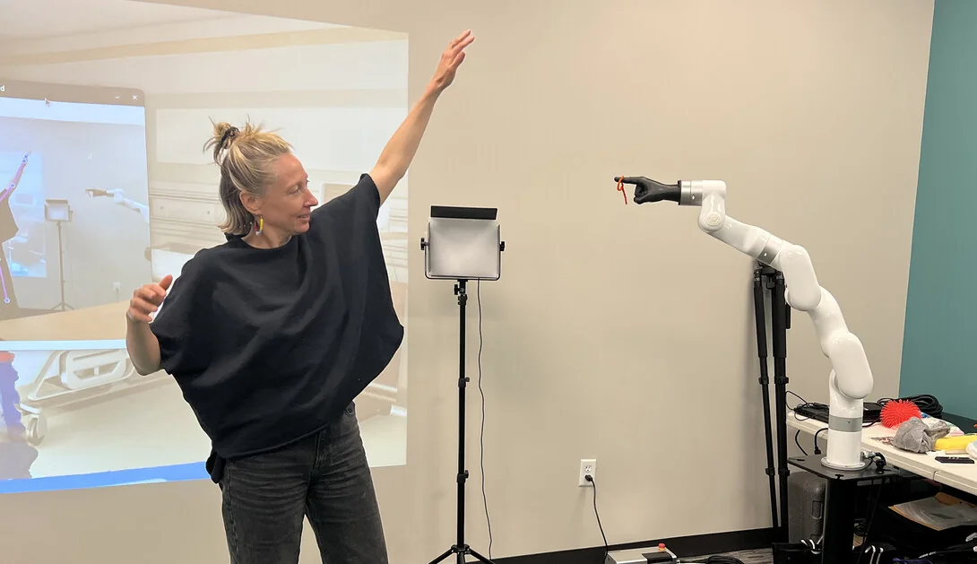 Ash Eliza Smith raises one arm and gestures with the other beside a white robotic arm, with a projected virtual care setting behind her.
